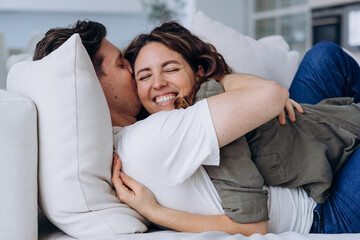Happy newlyweds with dark hair have fun tickling and hugging lying on sofa and kissing softly with love in living room extreme closeup - obrazy, fototapety, plakaty