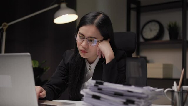 Asian businesswoman working with laptop computer in the dark office at night. Smart woman work hard overtime serious thinking hand on head. Feeling stressed and headache in warm light late at night