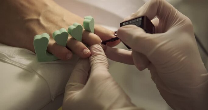 Pedicurist applying nail paint glaze to foot finger nails of woman client at parlor