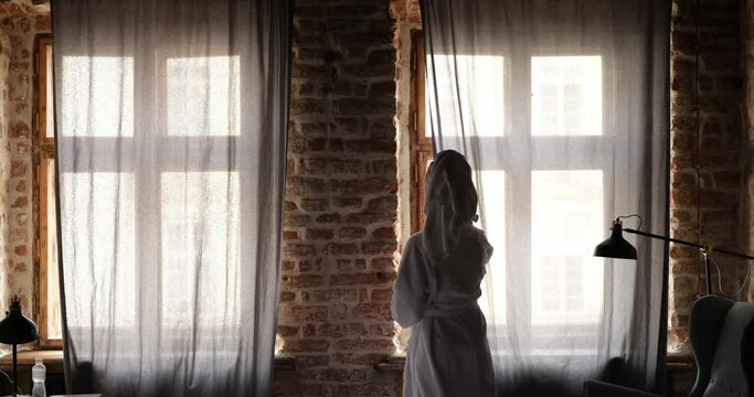Rear view of woman in bathrobe opening curtain and looking out through window