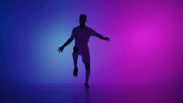 Black man a player juggles football ball in a dark studio with neon lights on the floor and red and blue lighting effects in slow motion. African professional football soccer player