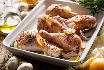 Raw chicken legs drumsticks with rosemary, garlic and lemon in a ceramic baking dish prepared for baking, close up view