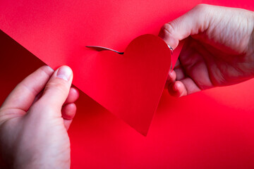 Male hands cutting a red heart from the paper with small scissors. Valentine's day DIY card theme.