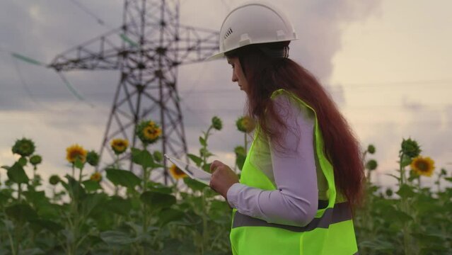 Green electricity. Power engineer woman in protective helmet maintains power line in sunflower field using data on tablet. Construction of high voltage power lines. Sale of electricity. Power supply