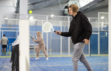 Teenage boy holding padel racquet in hand and ready to return ball while playing in court.