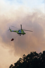 A helicopter flies by discharging water amid clouds of smoke from a forest fire