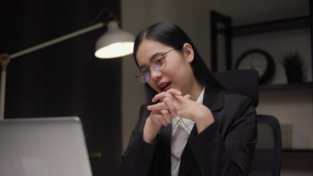 Young asian businesswoman working with laptop computer video conference in the dark office at night. Attractive Indian woman work hard overtime in home with floor lamp ambient warm light late at night