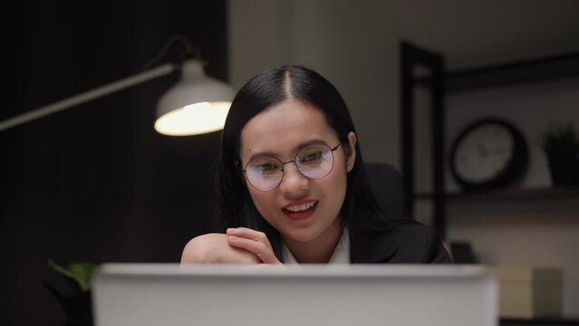 Young asian businesswoman working with laptop computer video conference in the dark office at night. Attractive Indian woman work hard overtime in home with floor lamp ambient warm light late at night
