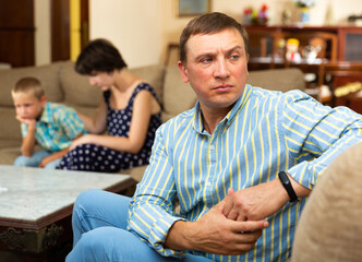 Unhappy man sitting at sofa after family quarrelling, woman with son on background