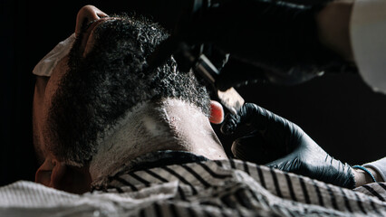 Fototapeta na wymiar Close-up of an unrecognizable man being shaved with razor on a black background, in a barber shop. Lifestyle.