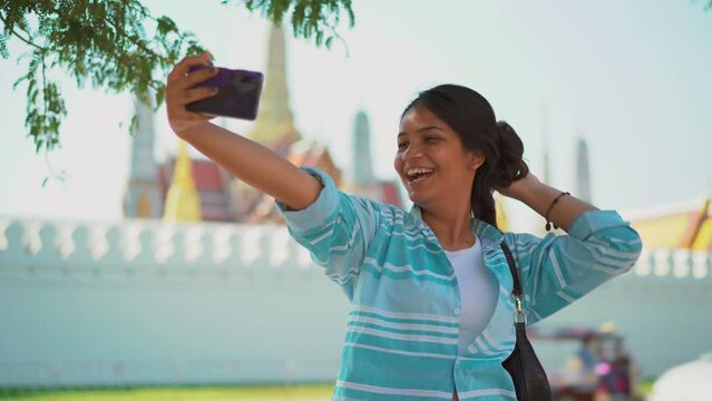 Beautiful joyful young tourist asian woman posing to smartphone camera and taking selfie photo at Thai Buddhist Temple. Pretty stylish female tourist making photos with mobile phone at cozy street