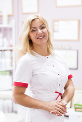 Fototapeta premium Portrait of blonde woman doctor in a beauty salon in white uniform, beautician looking at the camera and smiling, doctor stands on a background of certificates