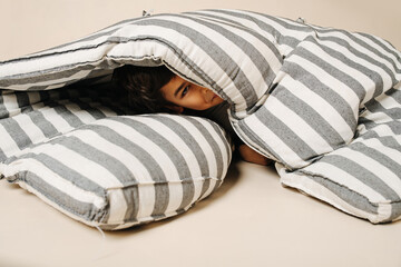 Playful indian boy lying under a striped folded mattress over beige background.