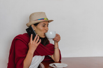 Latin peasant woman wearing hat and a red shawl seated drinking a cup of coffee while listening to...
