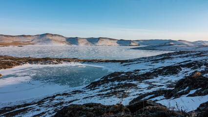 Two frozen lakes are located nearby. Snow on ice and on the ground. A mountain range against a blue sky. A sunny winter day. Baikal