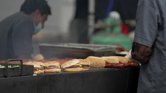 This close up video shows the arms of anonymous workers working on a bbq grill pit, cooking burgers and hot dogs.