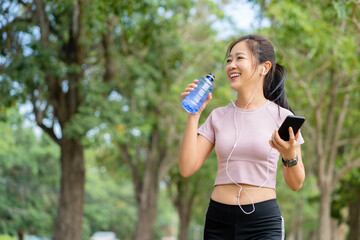 Young Woman drinking water after running and listening to music in the park. Running makes your body stronger. Concept of woman runners
