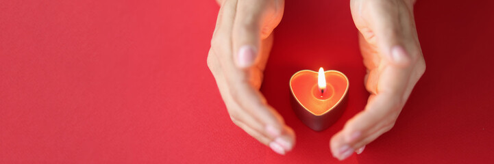 Female hand carefully protects burning candle in shape of heart on red background