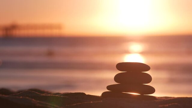 Stack of pebble stones, sandy ocean beach, sunset sky. Rock balancing in sun light, sea water waves. Stones staking in pyramid pile. Zen meditation and harmony in balance. Seamless looped cinemagraph.