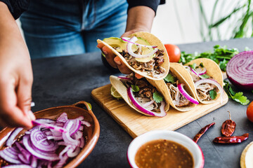 latin woman hands preparing mexican tacos with pork carnitas, avocado, onion, cilantro, and red sauce in Mexico