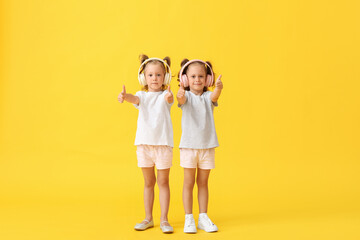 Cute little girls in headphones showing thumbs-up on yellow background