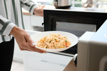 Woman putting plate of rice with vegetables into microwave oven in kitchen