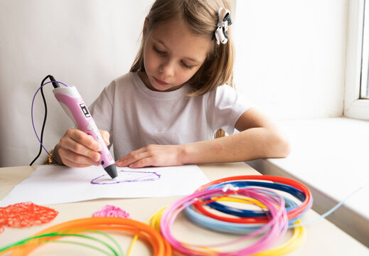 A 7-year-old girl with long hair and a white T-shirt is sitting at a table making figures with a pink 3-d pen. Toys made of plastic wire.