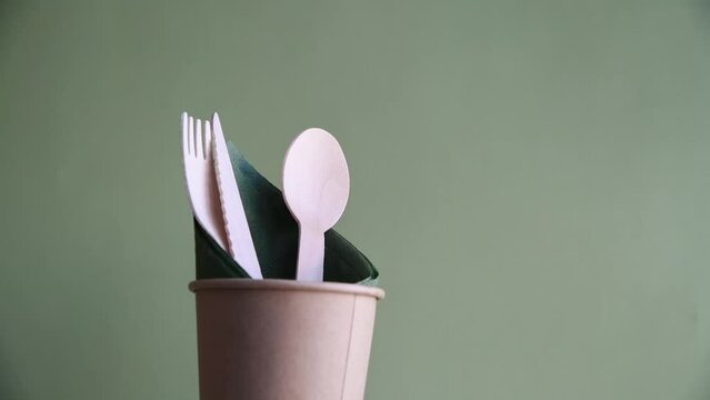Group of wooden disposable cutlery (forks, spoons and knifes) falls in brown paper cup with green napkin standing against green background. 4K resolution video. Environmental conservation theme.
