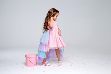 beautiful little girl in a chic pink dress poses standing on a gray background and holds a hatbox...