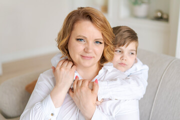 Obraz na płótnie Canvas Happy woman have fun with cute baby boy 5-6-7 years old in white shirt. Mom little baby son posing together hugs sitting on sofa in living room at home. Mothers day family love concept