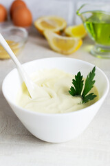Homemade mayonnaise in a white bowl with parsley leaf and ingredients in the background. The concept of food cooked at home. Selective focus. Vertical orientation.