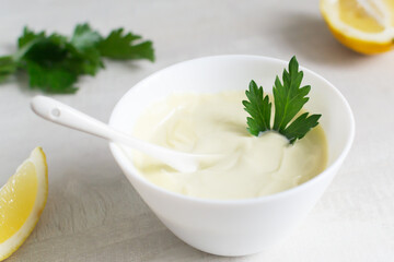 Homemade mayonnaise in a white bowl with parsley leaf and lemons in the background. The concept of food cooked at home. Selective focus. Vertical orientation.