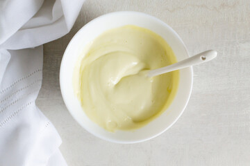 Homemade mayonnaise in a white bowl with a teaspoon and a white kitchen towel. Homemade food concept. Horizontal orientation. Top view.