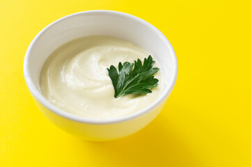 Homemade mayonnaise in a white bowl with a green parsley leaf on a yellow background. The concept of proper nutrition. Creative. Minimalism. Horizontal orientation. Copy space.