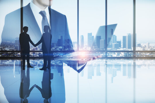 Abstract image of businessman using laptop computer in modern office interior with panoramic city view, businesspeople shaking hands and mock up place. Teamwork, leadership and technology concept. 