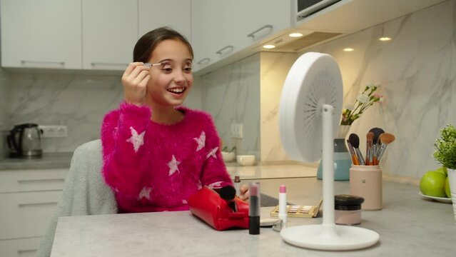 Adorable preteen girl applying eye makeup, looking at vanity mirror admiring her reflection indoors. Positive female child in pink pullover trying mom cosmetics, smiling charmingly, having fun at home