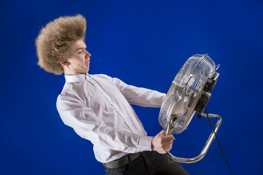 Funny Man. Fun. Humor. Hair Develops In The Wind. Strong Air Flow. A Man With Curly Hair Holds A Fan In His Hands. The Fan Blows The Guy Away. To Escape The Heat