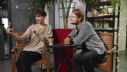 Two gay guys on a date are sitting in a coffee shop, drinking coffee and taking selfies on their phone. LGBT