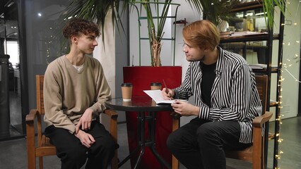 Two gay guys on a date are sitting in a coffee shop. The guy draws in his partner 's notebook. LGBT