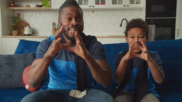 Excited African father and lovely school age boy watching American football live stream on tv, cheering team and celebrating successful reception with gesture and fist bump during super bowl match.