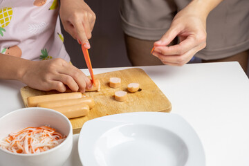 The hands of an Asian girl and his mother are cutting sausages to prepare a dinner menu.