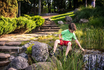 Naklejka na ściany i meble A young boy sits on the river bank, dreams. Warm summer or spring day. Cute Caucasian kid play with water
