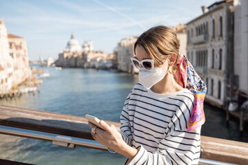 Fototapeta na wymiar Young woman in medical mask traveling during pandemic in Italy. Concept of social rules of wearing a mask during a pandemic. Woman using smartphone while standing on the bridge in Venice