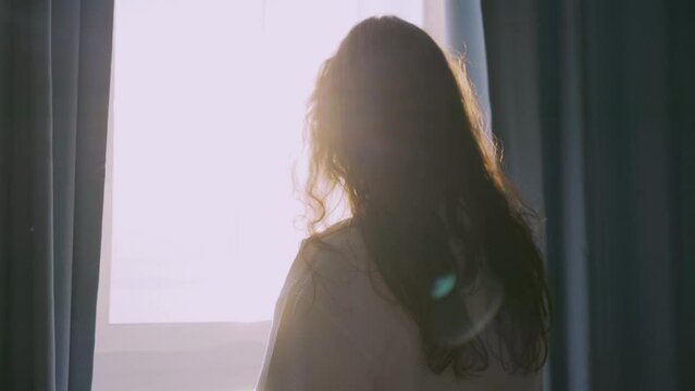 Curly haired lady in white bathrobe opens curtains in dark hotel room. Backside woman silhouette to look outside large window at sunrise time, closeup