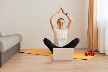 Online sport fitness yoga training workout. Young woman doing exercises on yoga mat opposite laptop at home, raised arms, keeps palms together, wearing white T-shirt and black leggins .