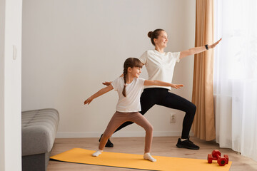 Fototapeta premium Indoor shot of beautiful young woman and her charming little daughter are smiling while doing yoga together at home, standing on yoga mat, doing warrior pose.