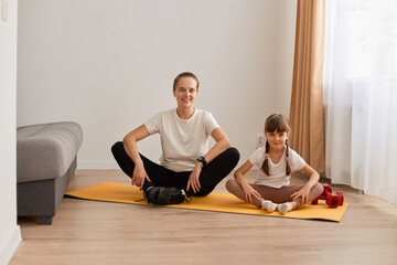 Portrait of smiling peaceful woman with hair bun sitting on mat practicing yoga with her daughter, meditating and relaxing together with child, looking at camera with happy expression.