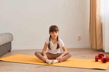 Portrait of cute little girl with pigtails sitting on fitness mat in a lotus position while doing yoga at home, meditating playing sports, looking at camera with calm expression.