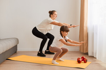 Mother and little child daughter practicing are engaged in fitness, sport exercise mat at home, woman with child doing squatting together with outstretched hands.