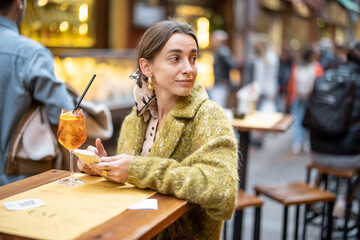 Woman sitting with a cocktail and phone at outdoor bar or restaurant on the crowded street in Bologna. Concept of Italian lifestyle and gastronomy